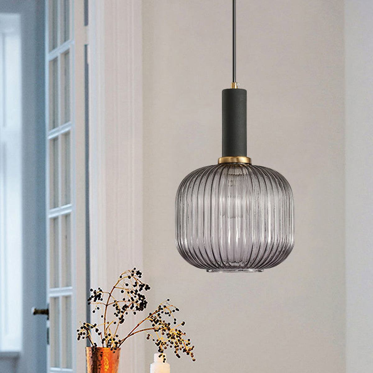 Rounded smoke grey reeded glass pendant from Clérachic with a black fitting hangs by a white-framed window with dried berry stems below.