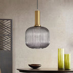 Rounded smoke grey reeded glass pendant from Clérachic on a gold stem hangs against a stone-effect wall, with green vases and a dark bowl.