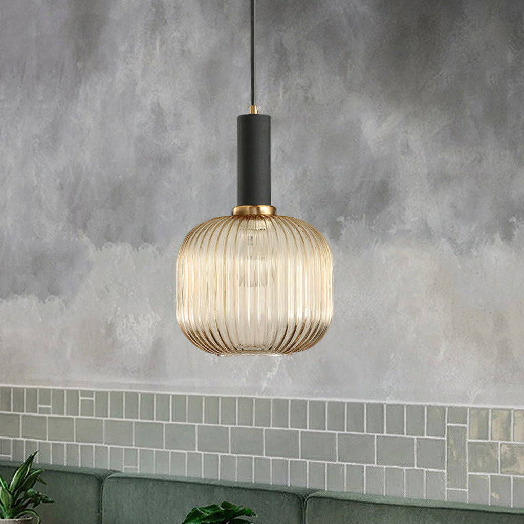 A lit, rounded amber reeded glass pendant from Clérachic with a black fitting, hanging against a grey wall above green wall tiles.