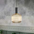 A lit, rounded amber reeded glass pendant from Clérachic with a black fitting, hanging against a grey wall above green wall tiles.