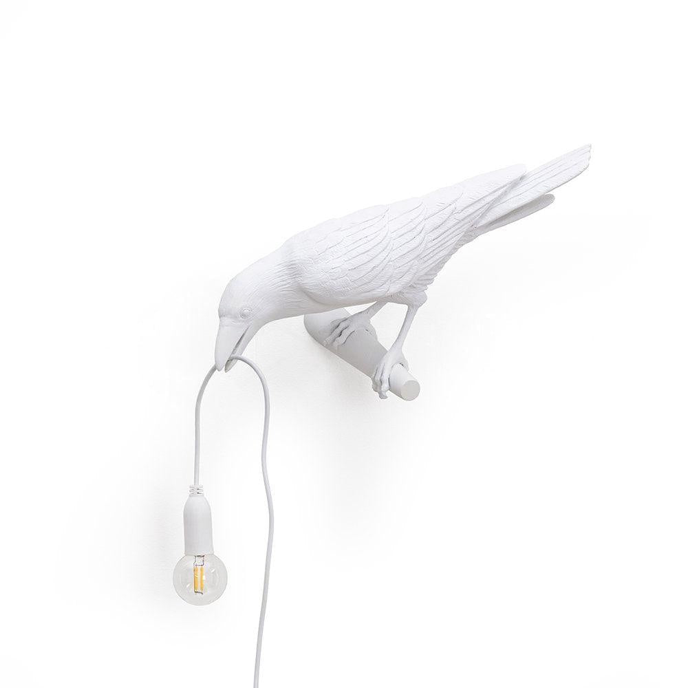 From another angle, this white Clérachic bird lamp shows its wall mount clearly, bulb glowing warm on plain white.