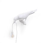 From another angle, this white Clérachic bird lamp shows its wall mount clearly, bulb glowing warm on plain white.