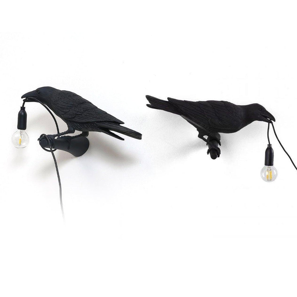 Two black Clérachic bird lamps stand side by side, each gripping its own hanging bulb and cord in its beak.