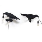Two black Clérachic bird lamps stand side by side, each gripping its own hanging bulb and cord in its beak.