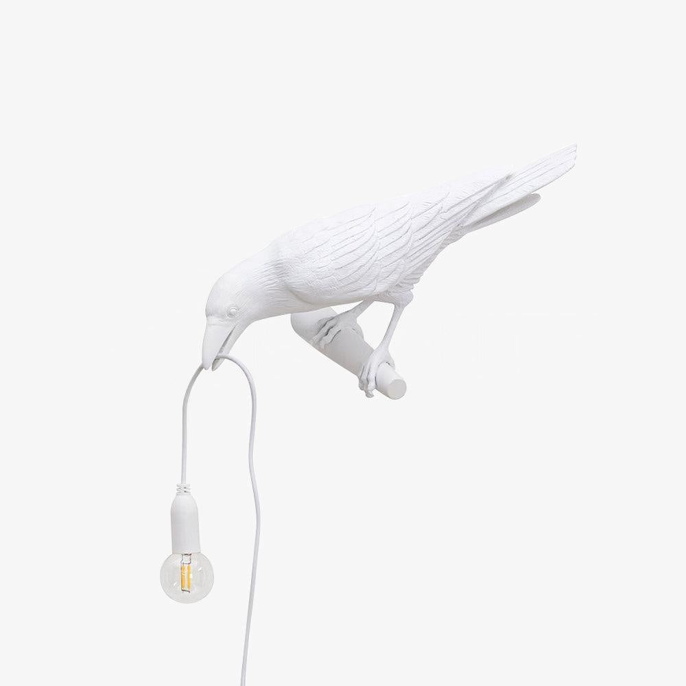 Isolated on white, this white resin Clérachic bird lamp bends down with its beak on the cord, bulb glowing warm.