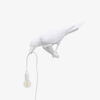 Isolated on white, this white resin Clérachic bird lamp bends down with its beak on the cord, bulb glowing warm.
