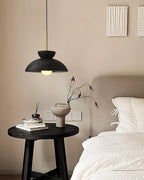Small black dome pendant from Clérachic above a bedside table with a vase and books, beside a made-up bed.