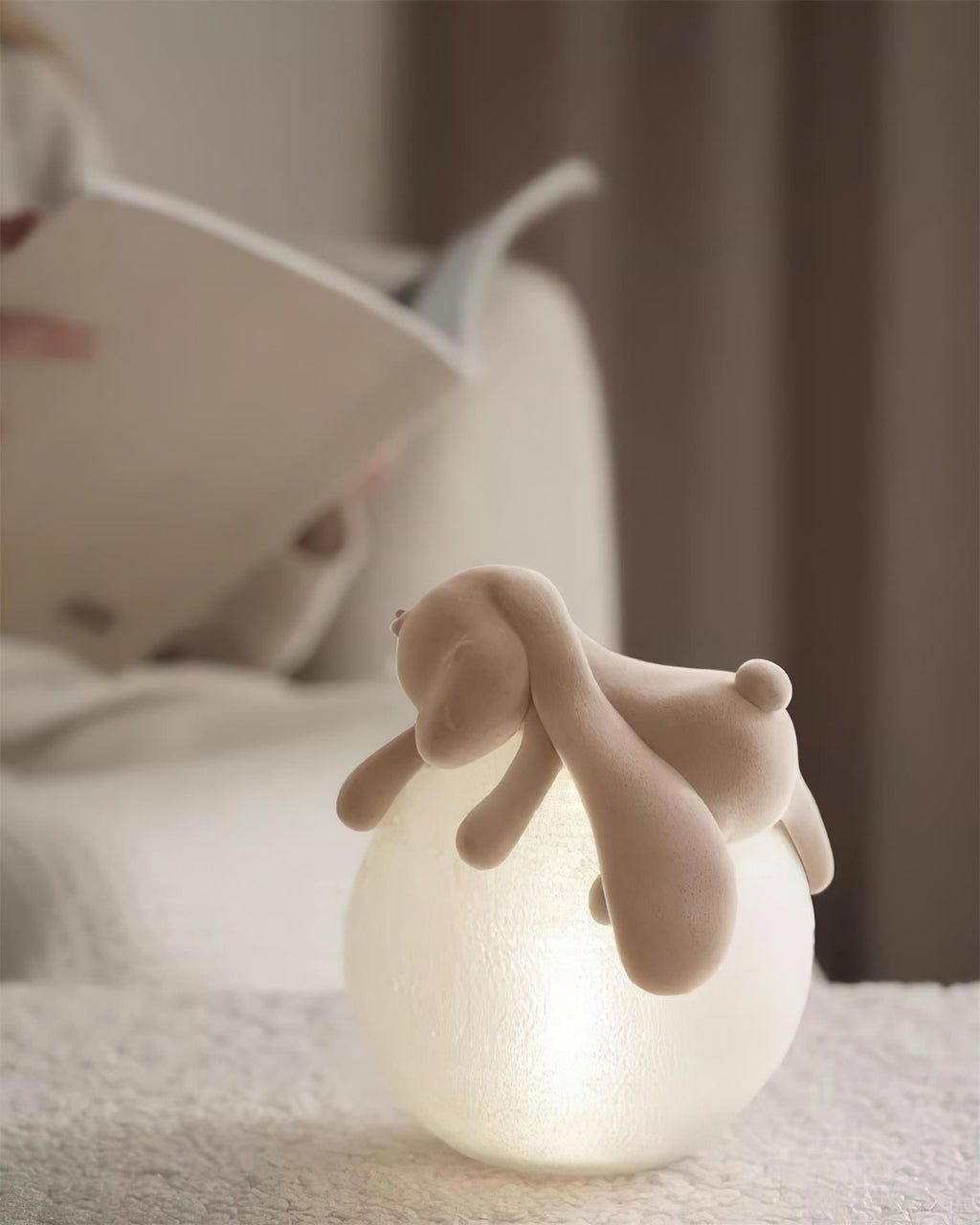 Lit softly, a Clérachic rabbit table lamp sits on a cream boucle surface, an open book behind it.