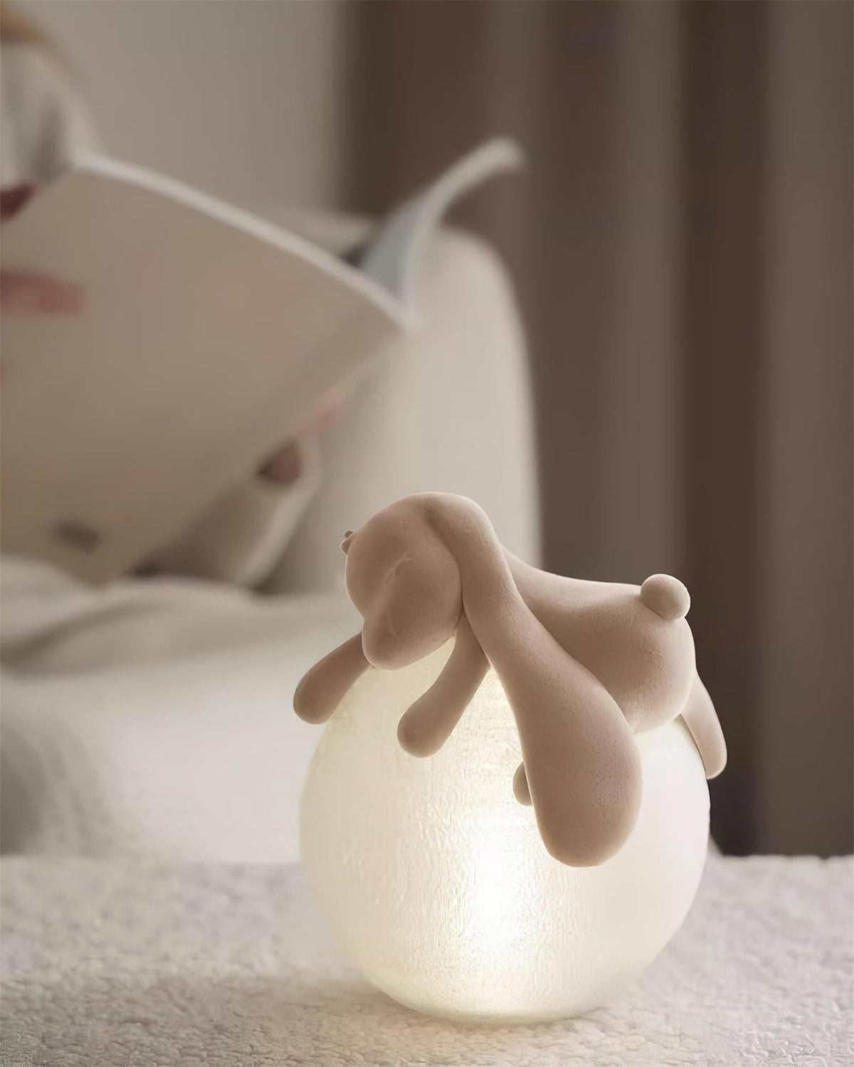 Lit softly, a Clérachic rabbit table lamp sits on a cream boucle surface, an open book behind it.