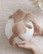 Held in two hands, a Clérachic rabbit lamp shows its flocked body and pale textured globe, above a cream boucle surface.