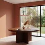 A Clérachic orange disc pendant hanging over a round walnut dining table, with a tall window and terracotta walls behind.