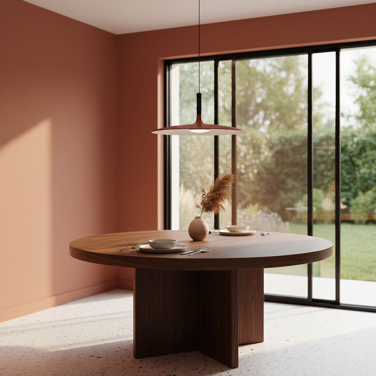 A Clérachic orange disc pendant hanging over a round walnut dining table, with a tall window and terracotta walls behind.