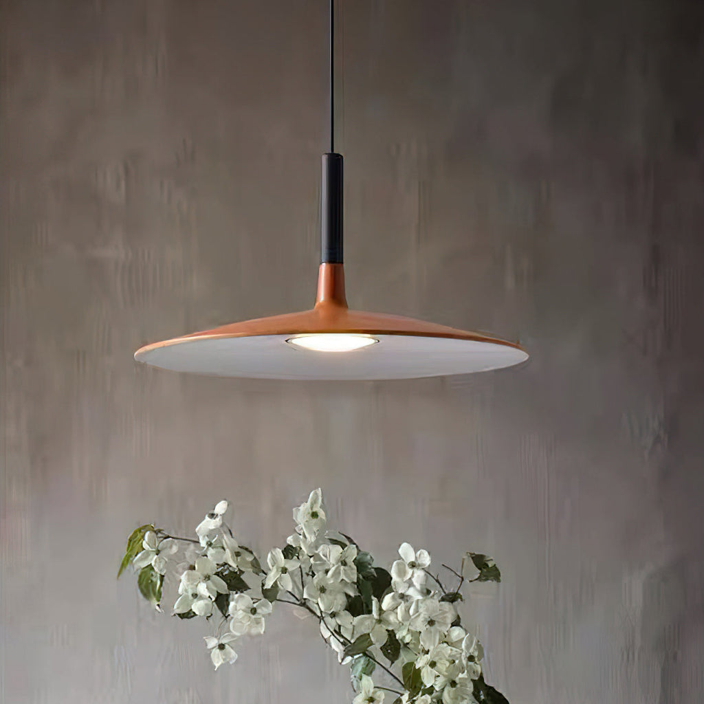 Close view of the wide orange disc shade and black stem of this Clérachic pendant, with white blossom below and a grey wall.