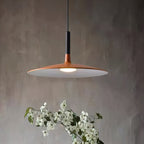Close view of the wide orange disc shade and black stem of this Clérachic pendant, with white blossom below and a grey wall.