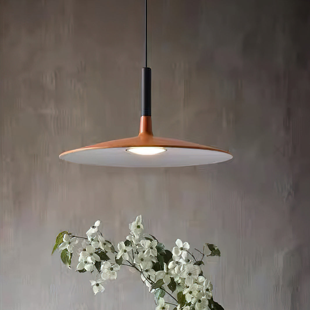 Close view of the wide orange disc shade and black stem of this Clérachic pendant, with white blossom below and a grey wall.