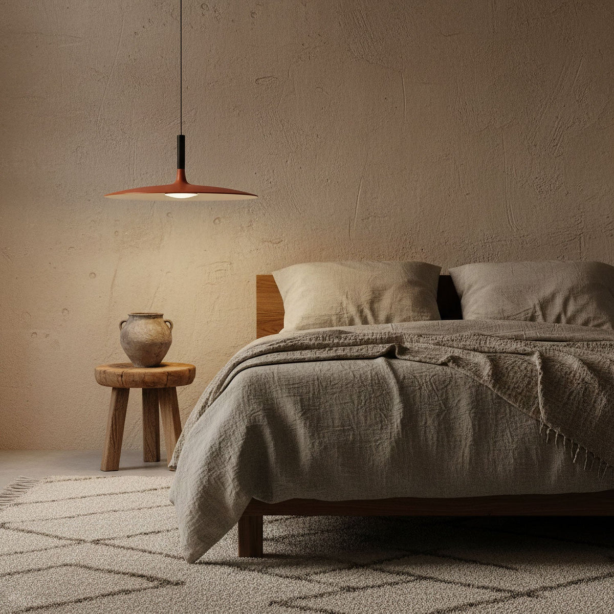 A Clérachic orange pendant light over a bed with beige linen, beside a small round wooden stool in a warm plaster-toned room.