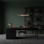 Clérachic disc pendant light in a dark green study, hanging above a black desk with a small lamp and a bookcase behind.