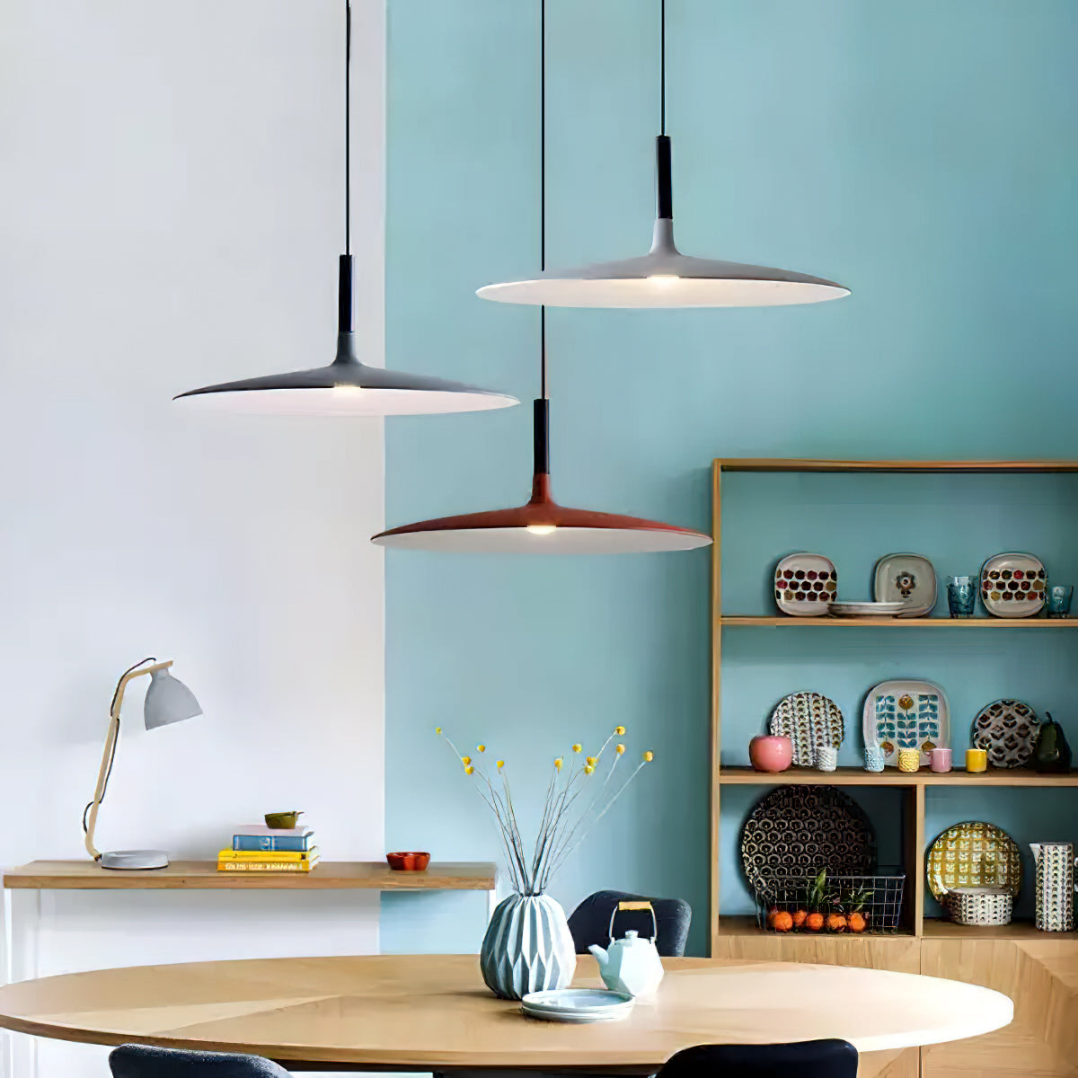 Three Clérachic disc pendants in black, grey and orange at different heights, against a pale blue wall and shelf of ceramics.