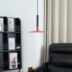 Red-orange Clérachic pendant light beside a pale curtain and a black shelf of magazines, with a leather chair at the edge.