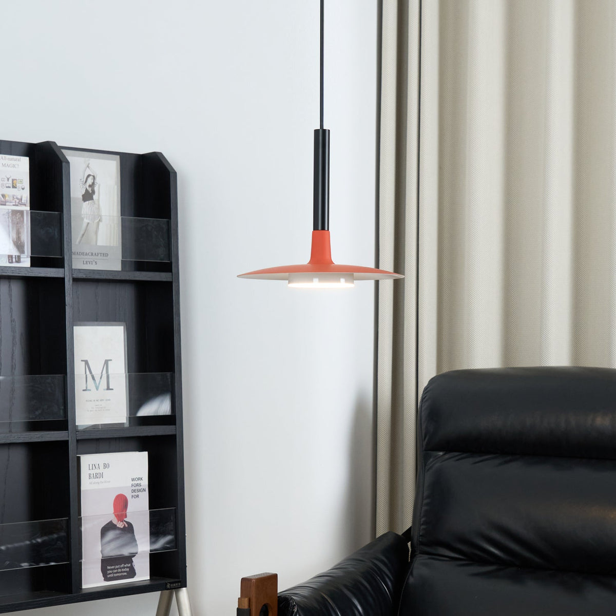 Red-orange Clérachic pendant light beside a pale curtain and a black shelf of magazines, with a leather chair at the edge.