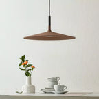 Brown Clérachic disc pendant on a pale wall, above a small vase of orange flowers and a stack of white cups.