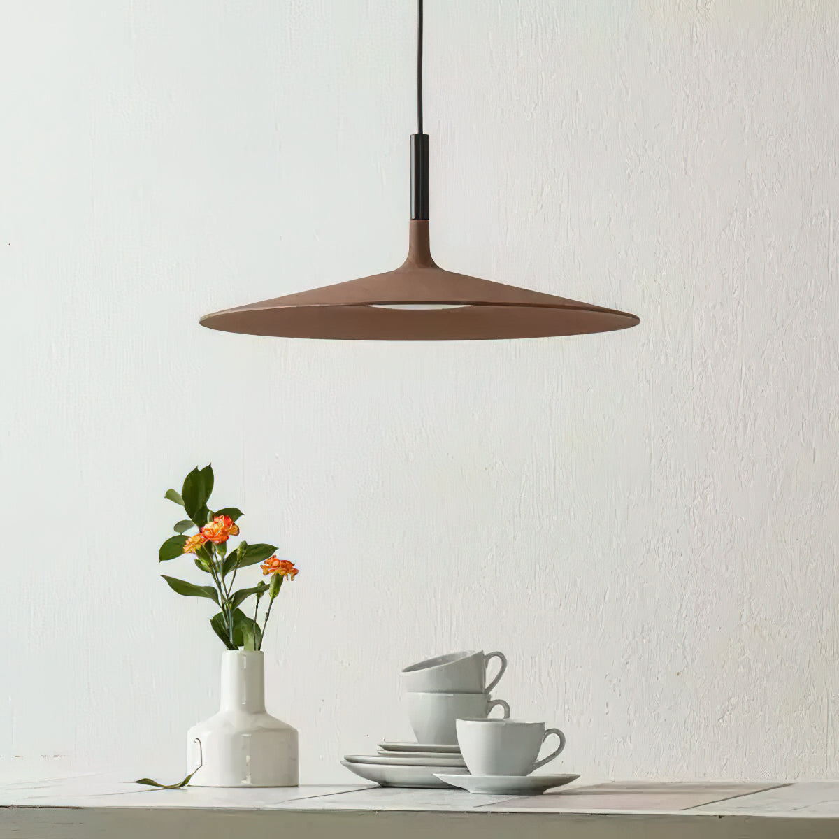 Brown Clérachic disc pendant on a pale wall, above a small vase of orange flowers and a stack of white cups.