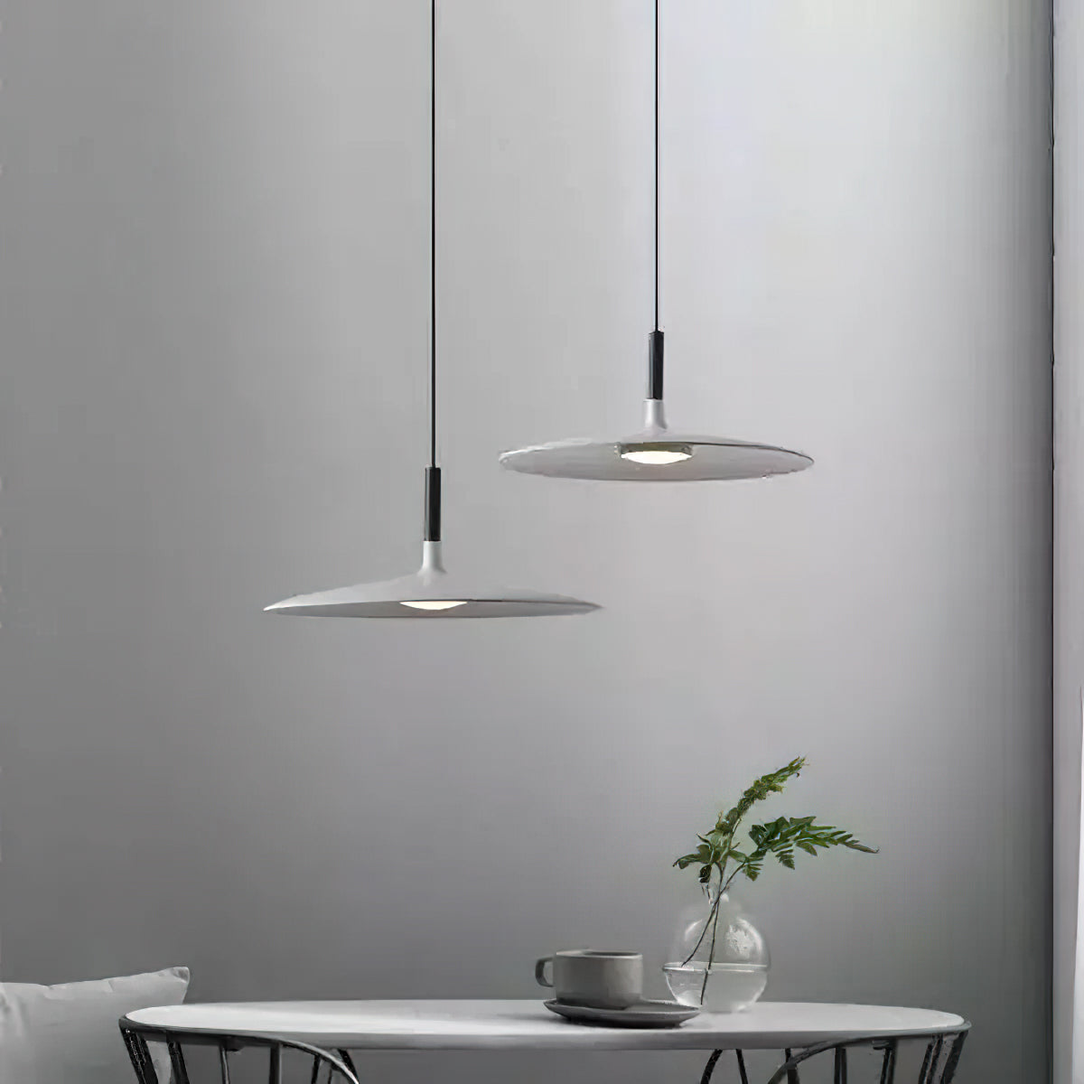Two Clérachic grey disc pendants at different heights on a pale grey wall, above a table with a cup and a leafy sprig.
