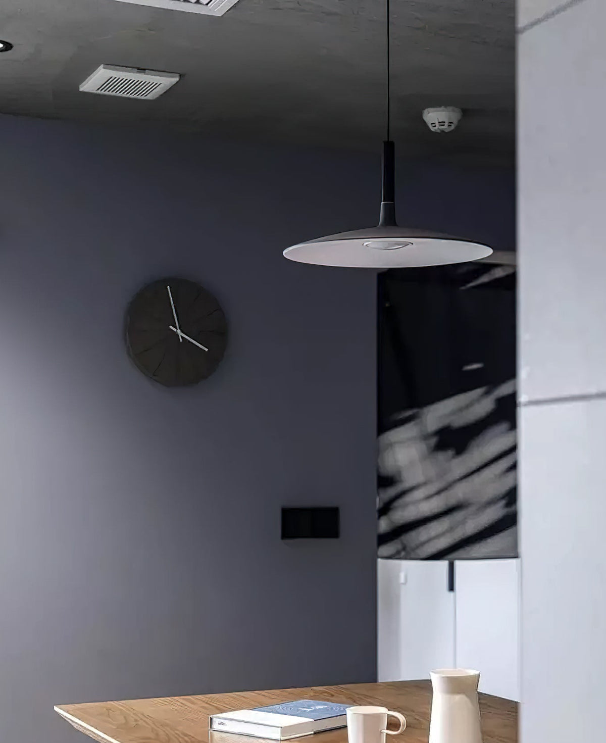 A grey Clérachic pendant light in a room with a dark blue-grey wall, a round clock and a table with a notebook below.