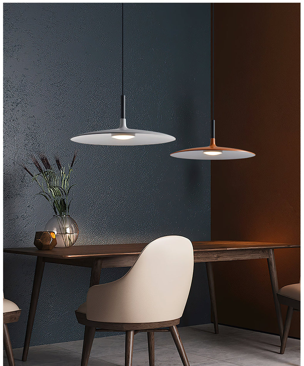 A grey and an orange Clérachic pendant over a dark wood dining table, with a cream chair and a glass vase of grasses.