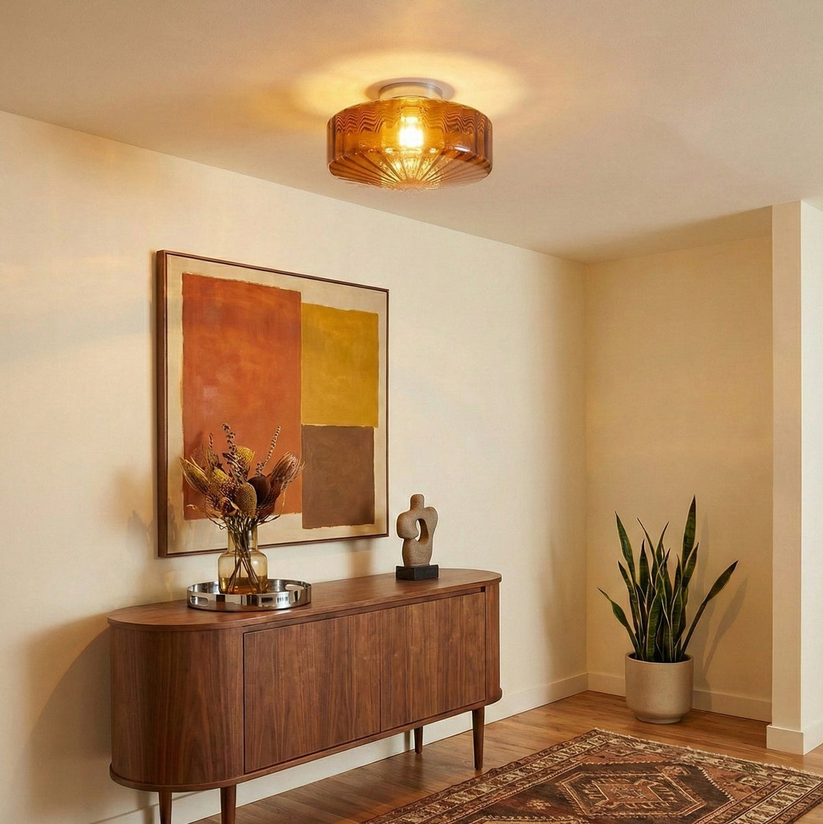 A lit brown glass Clérachic ceiling light above a curved walnut sideboard, with a dried-flower vase and a potted plant nearby.