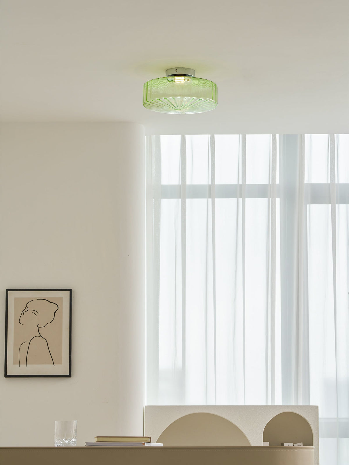 A small green Clérachic ceiling light on a white ceiling beside tall sheer curtains, with a framed line drawing on the wall.