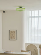 Distant view of a green Clérachic ceiling light high on a white ceiling, above a framed print and a desk.