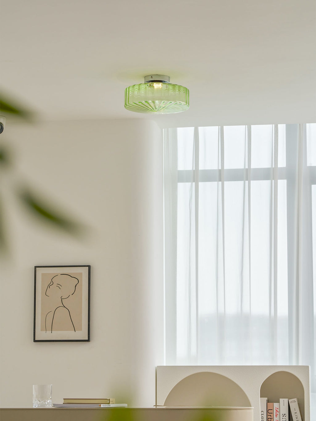 A green ribbed glass Clérachic ceiling light with a blurred leaf in the foreground, above a window and a framed print.
