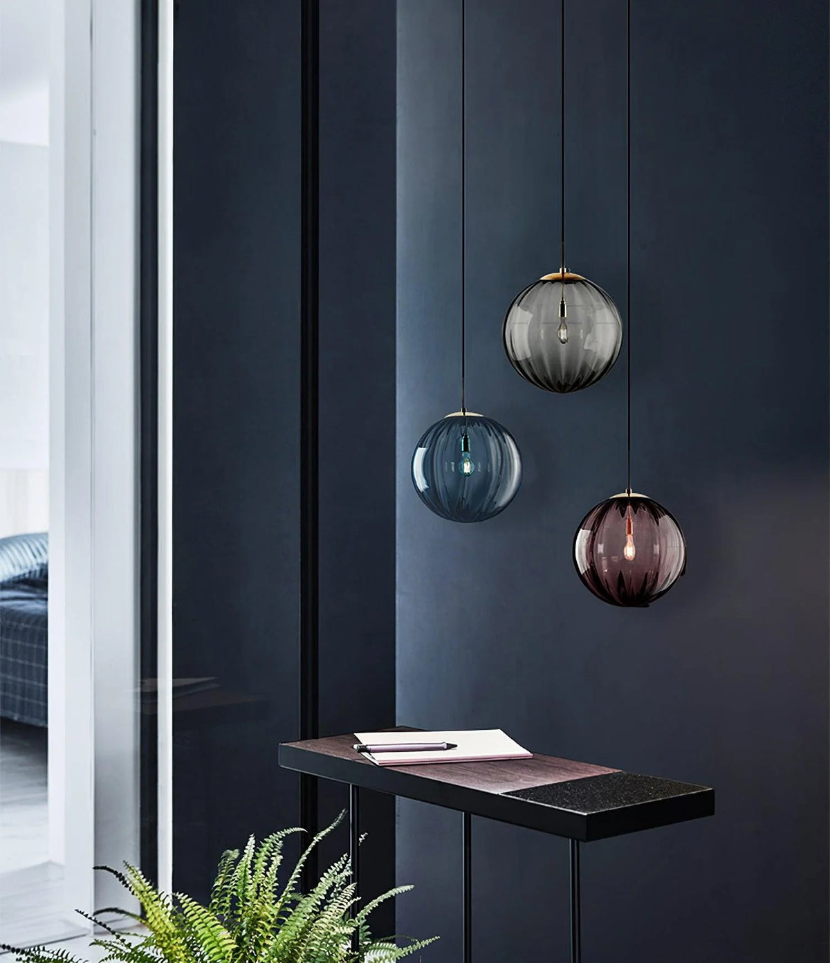 Three Clérachic glass globes at different heights in smoke grey, blue and wine red against a dark navy wall.