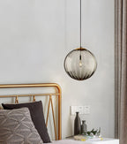 Smoke grey ribbed glass Clérachic pendant hung close to a gold bed frame and a beige curtain.