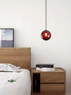Small wine red Clérachic glass pendant hanging by a wooden bed, with a bedside cabinet and books.