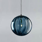 Large blue Clérachic glass globe with vertical ribs and a black cap, a bulb lit inside on a grey backdrop.