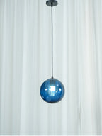 Small blue Clérachic glass pendant on a black cord against a sheer white curtain.
