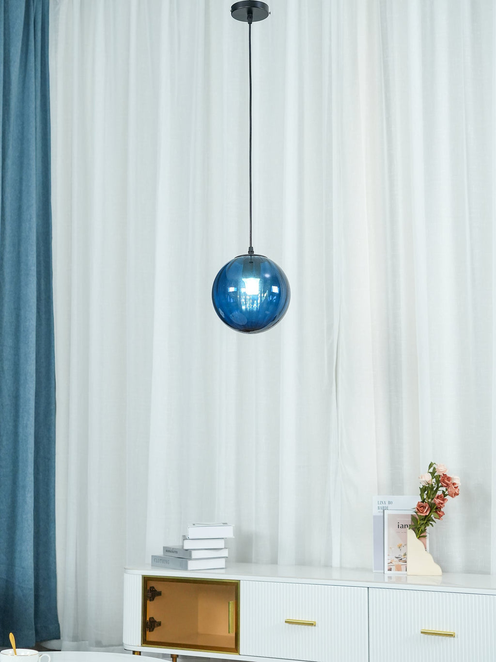 Blue Clérachic glass pendant against sheer curtains, a white sideboard with books and flowers below.