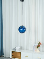 Dark blue Clérachic glass pendant unlit in front of white curtains and a sideboard with a vase of flowers.