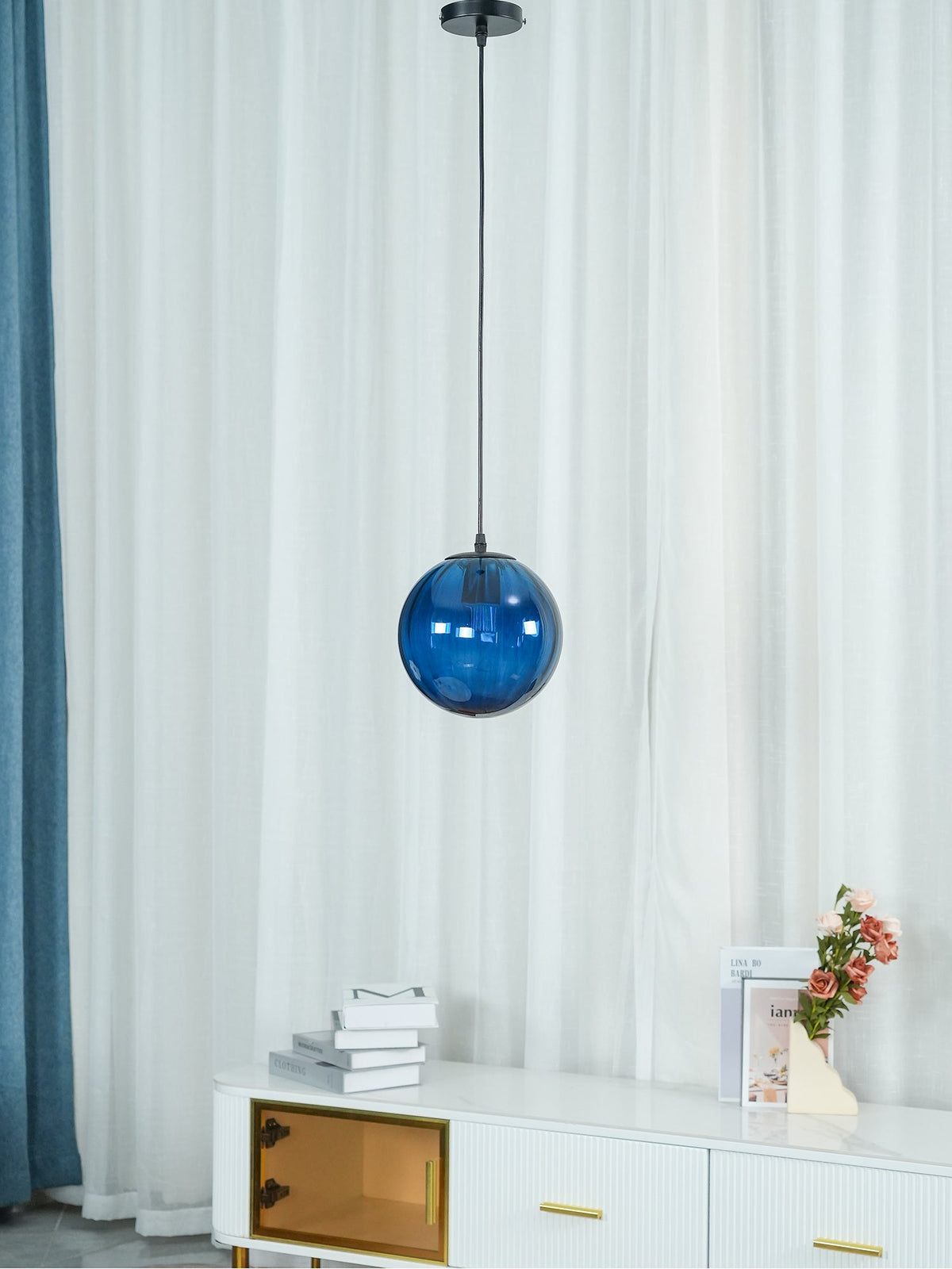Dark blue Clérachic glass pendant unlit in front of white curtains and a sideboard with a vase of flowers.