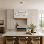 A Clérachic pendant ring above a white kitchen island with woven wooden stools, cream cabinets and a brass-edged hood.
