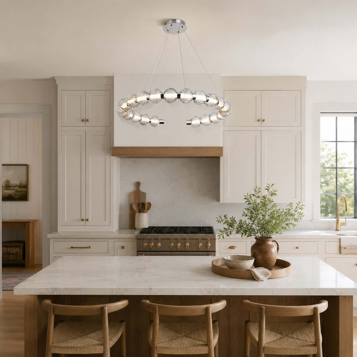 A Clérachic pendant ring above a white kitchen island with woven wooden stools, cream cabinets and a brass-edged hood.