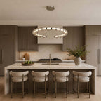This Clérachic pendant hangs over a long stone island with four cream bar stools, in a kitchen of warm brown cabinets.