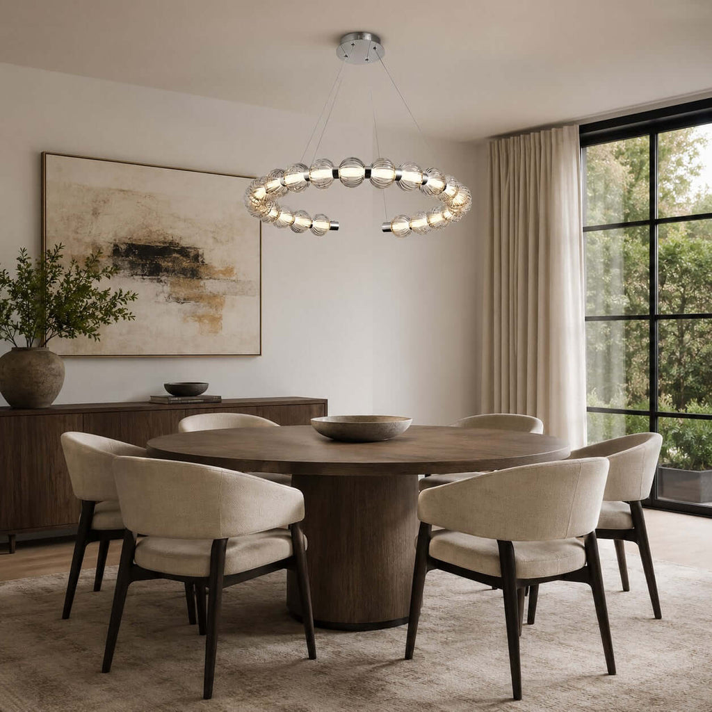 A Clérachic pendant ring over a round wooden dining table with four cream chairs, beside a tall black-framed window.