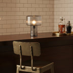 Lit ribbed glass Clérachic table lamp on a dark wood counter, in front of cream metro tiles, with a decanter and cocktail shaker.
