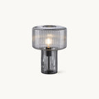 A ribbed smoky grey glass Clérachic table lamp with a wide drum shade over a slim fluted base, lit, on a plain white backdrop.