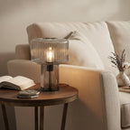 A smoky grey ribbed glass Clérachic lamp glows on a round side table beside a cream sofa, an open book and a lavender sprig.