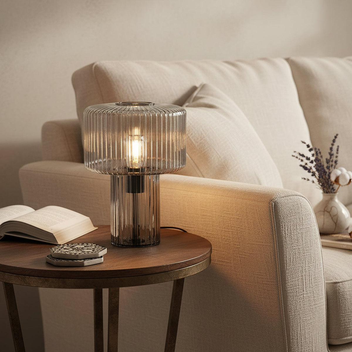 A smoky grey ribbed glass Clérachic lamp glows on a round side table beside a cream sofa, an open book and a lavender sprig.