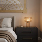 Warm-lit cognac Clérachic table lamp on a dark bedside chest with a marble top, below a gold fan-shaped mirror.